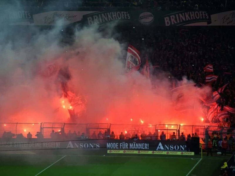 Tribune de football en feu avec des supporters brandissant des fumigènes rouges lors d'un match.